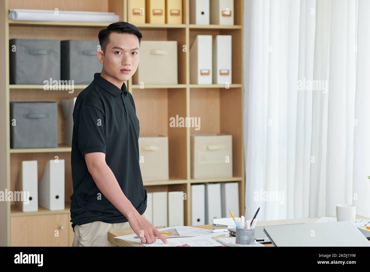 Serious young architecture bureau worker standing at desk with documents and blueprints Stock Photo