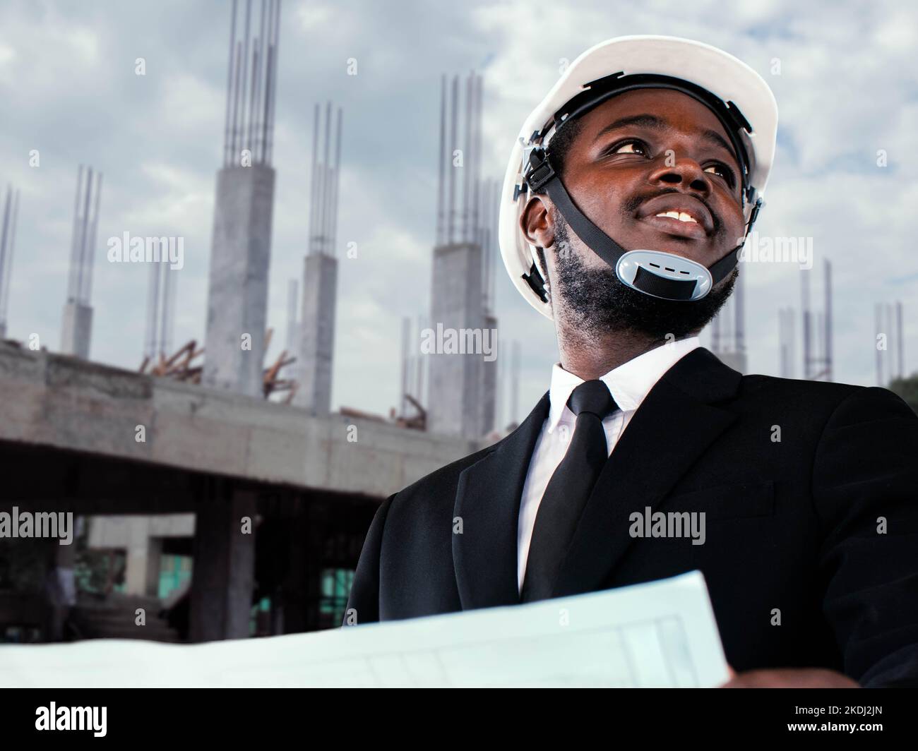 African manager engineer in suit wears a helmet and carries blue printed paper to inspect building construction within the construction zone Stock Photo