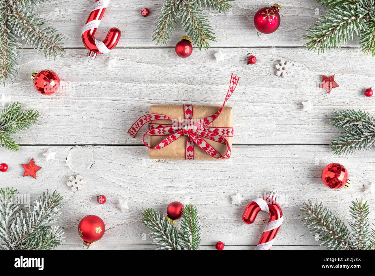 Christmas or Happy New Year background. Gift box in frame made of fir tree branches, red holiday decorations on white wooden background. Flat lay. Top Stock Photo