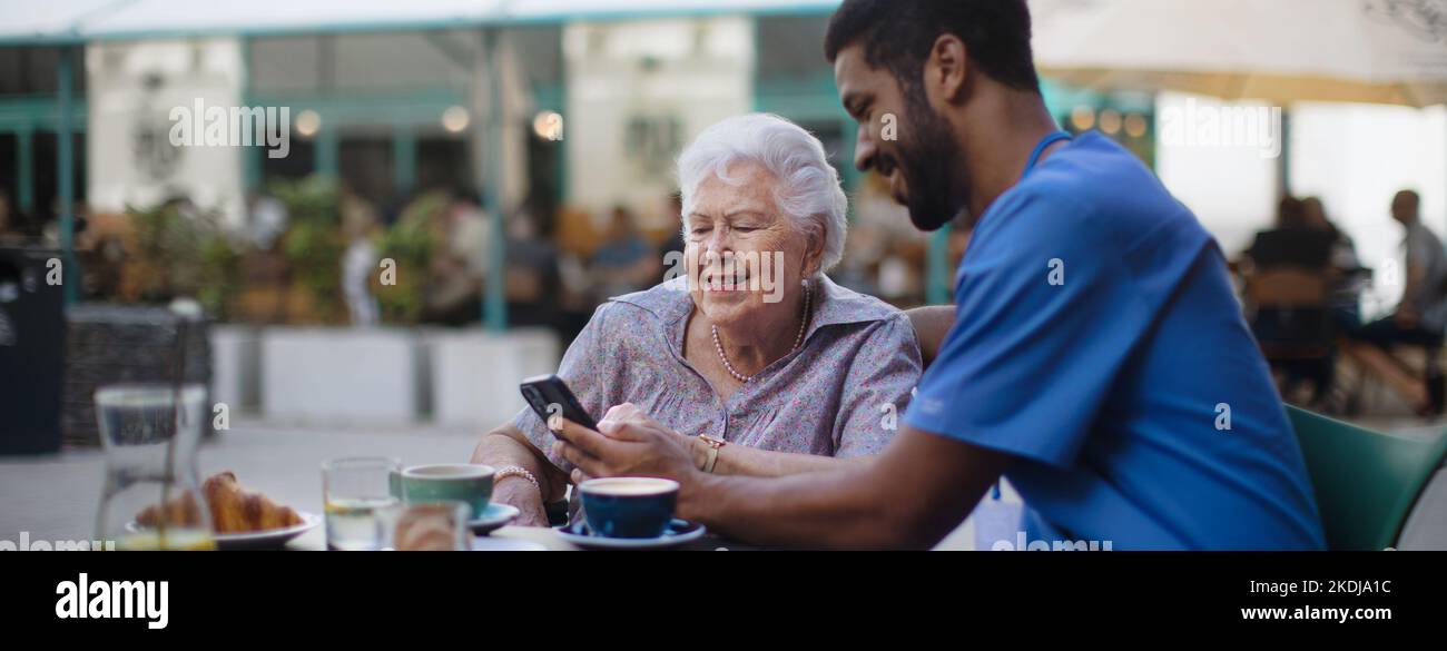 Caregiver having coffee with his client and learning her using smartphone, outdoor at cafe. Stock Photo