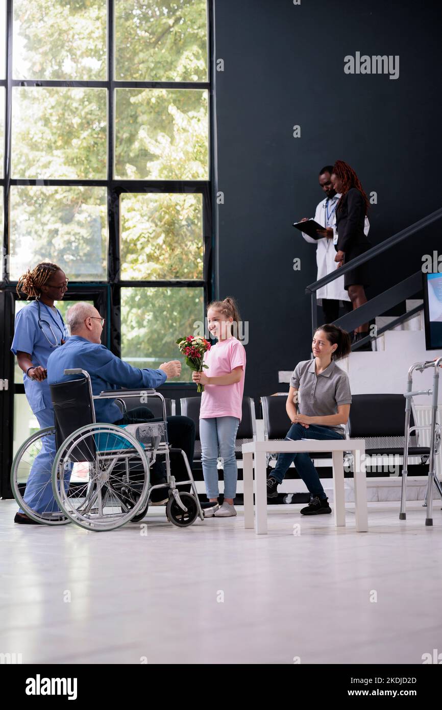 Little girl giving bouquet of flowers to disabled grandfather during checkup visit appointment in hospital waiting room. Patient in wheelchair discussing health care treatment with medical assistant Stock Photo