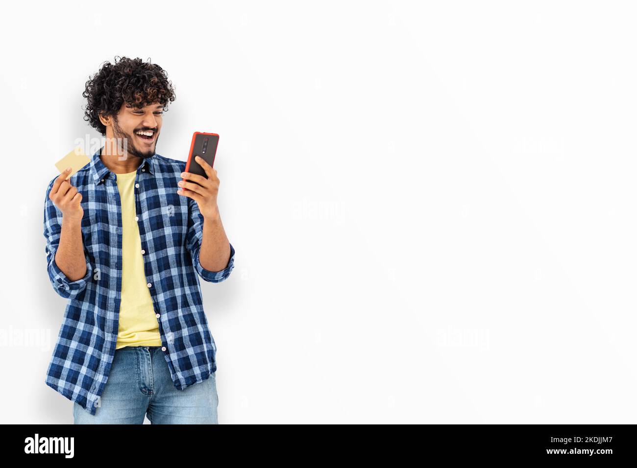 Smiling happy Asian man with curly hair holding mobile phone and credit card standing on white background Stock Photo