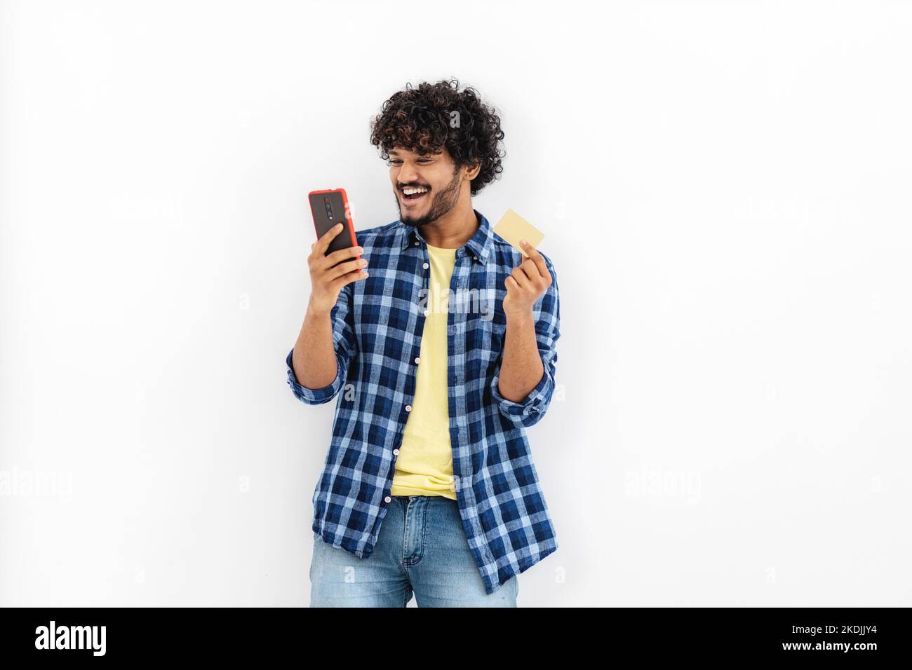 Smiling happy Asian man with curly hair holding mobile phone and gold credit card standing on white background Stock Photo