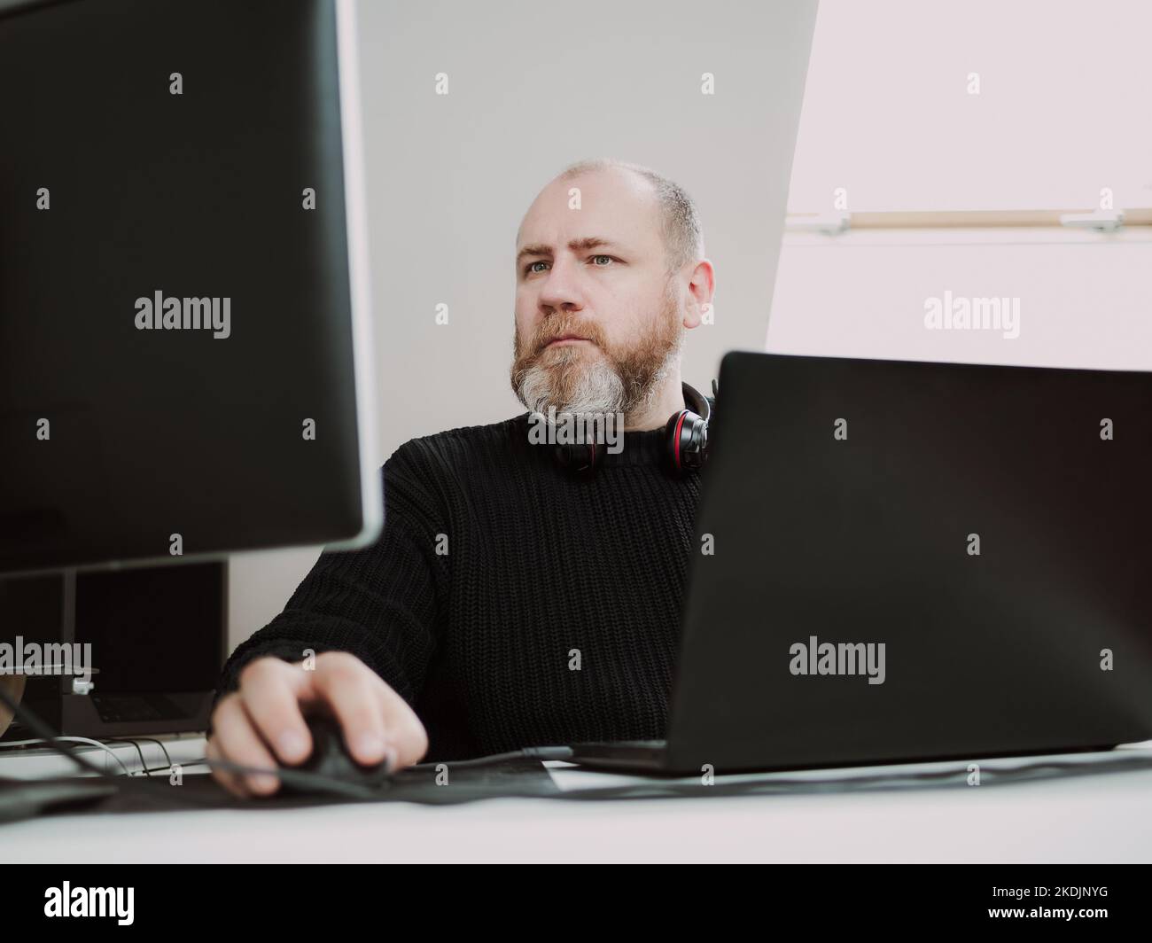 Adult focused bearded man remote working on laptop and using two monitors from home Stock Photo