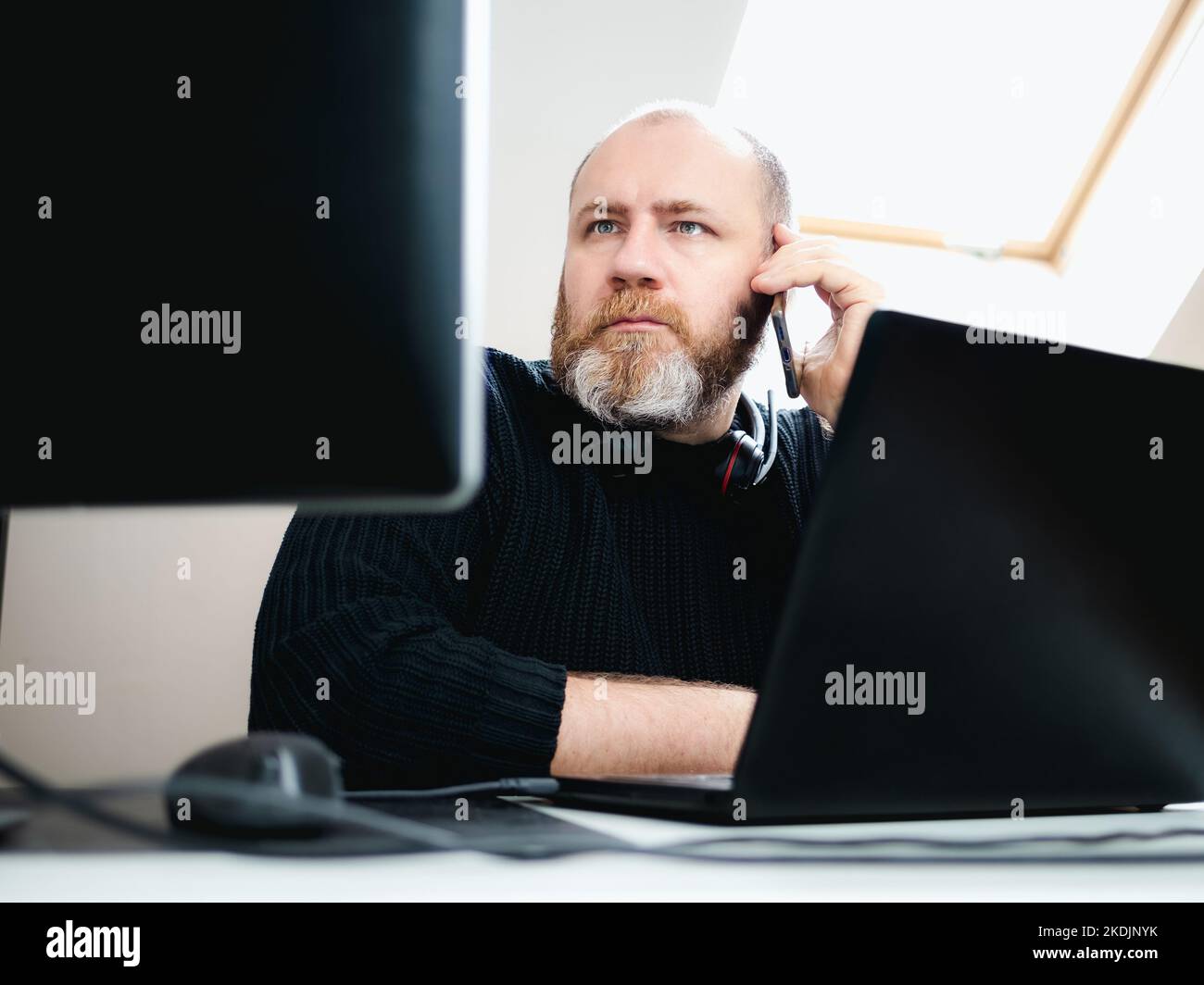 Adult focused man calling by smartphone during remote work on laptop and using two monitors from home Stock Photo