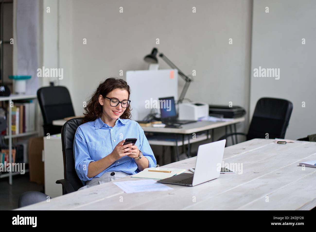 Smiling young female worker using cellphone working in office sitting at desk. Stock Photo