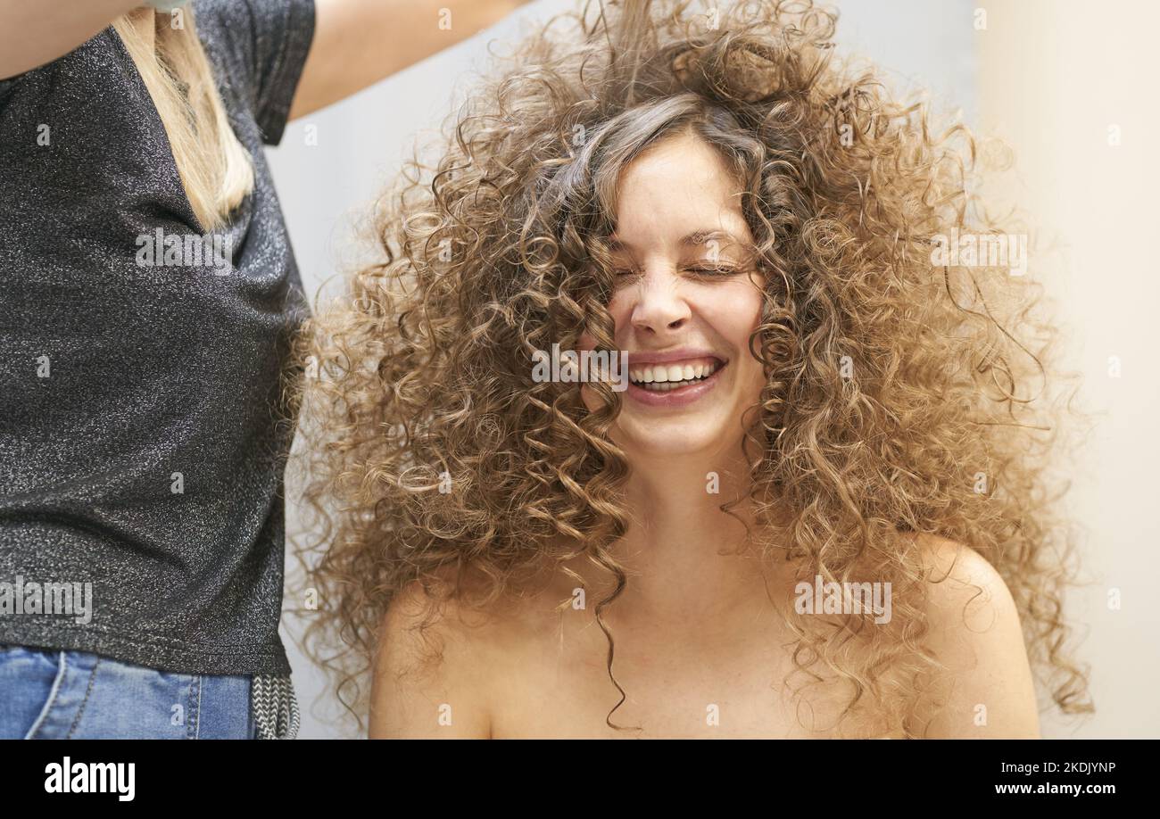 The hair master in face mask makes a curly hairstyle for a smiling woman. Portrait of a beautiful Caucasian girl with flowing curly hair. The concept of beauty salon. High quality photo Stock Photo