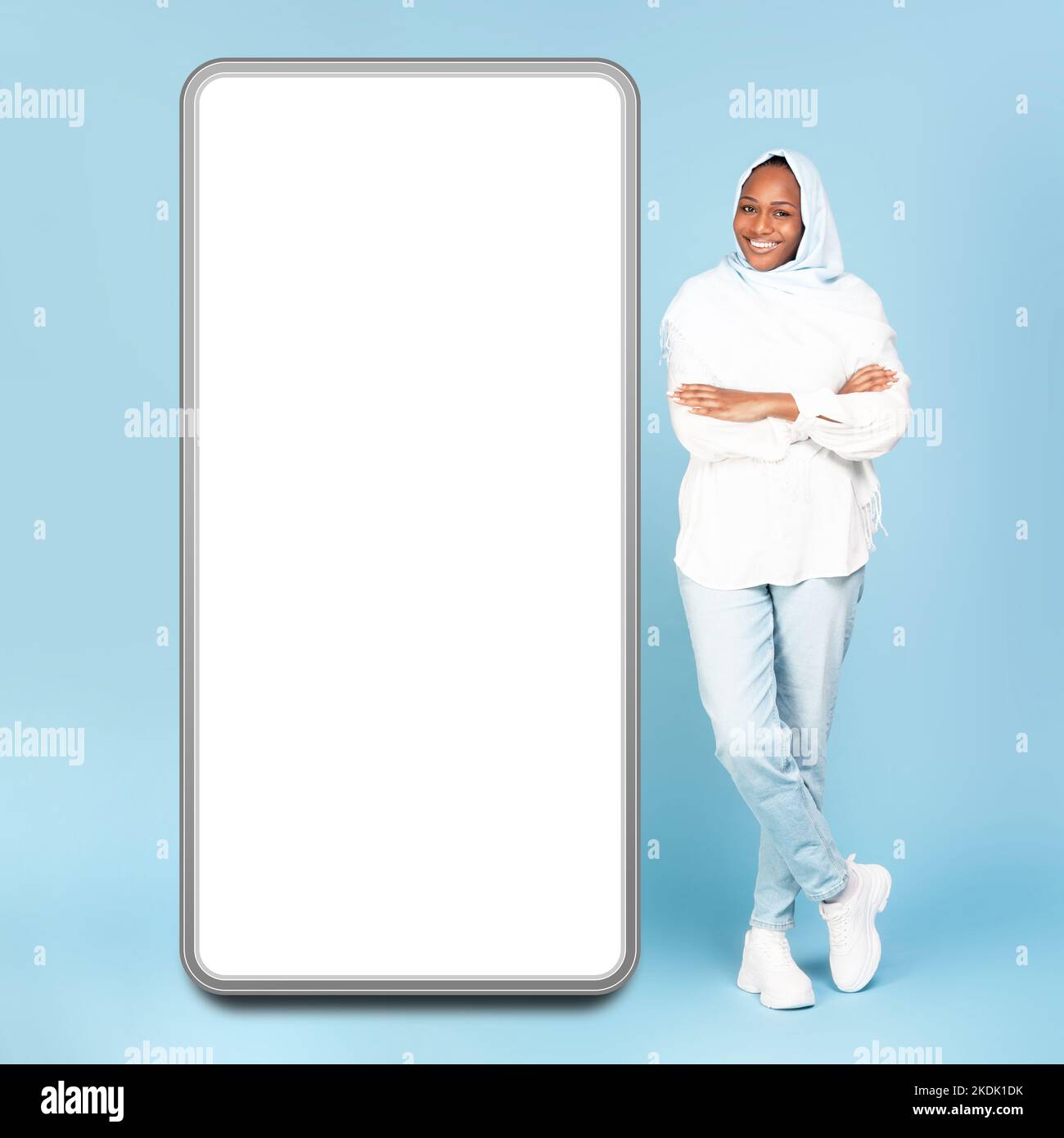Full body length of happy black muslim woman standing with folded arms near giant cellphone, empty screen, mockup Stock Photo