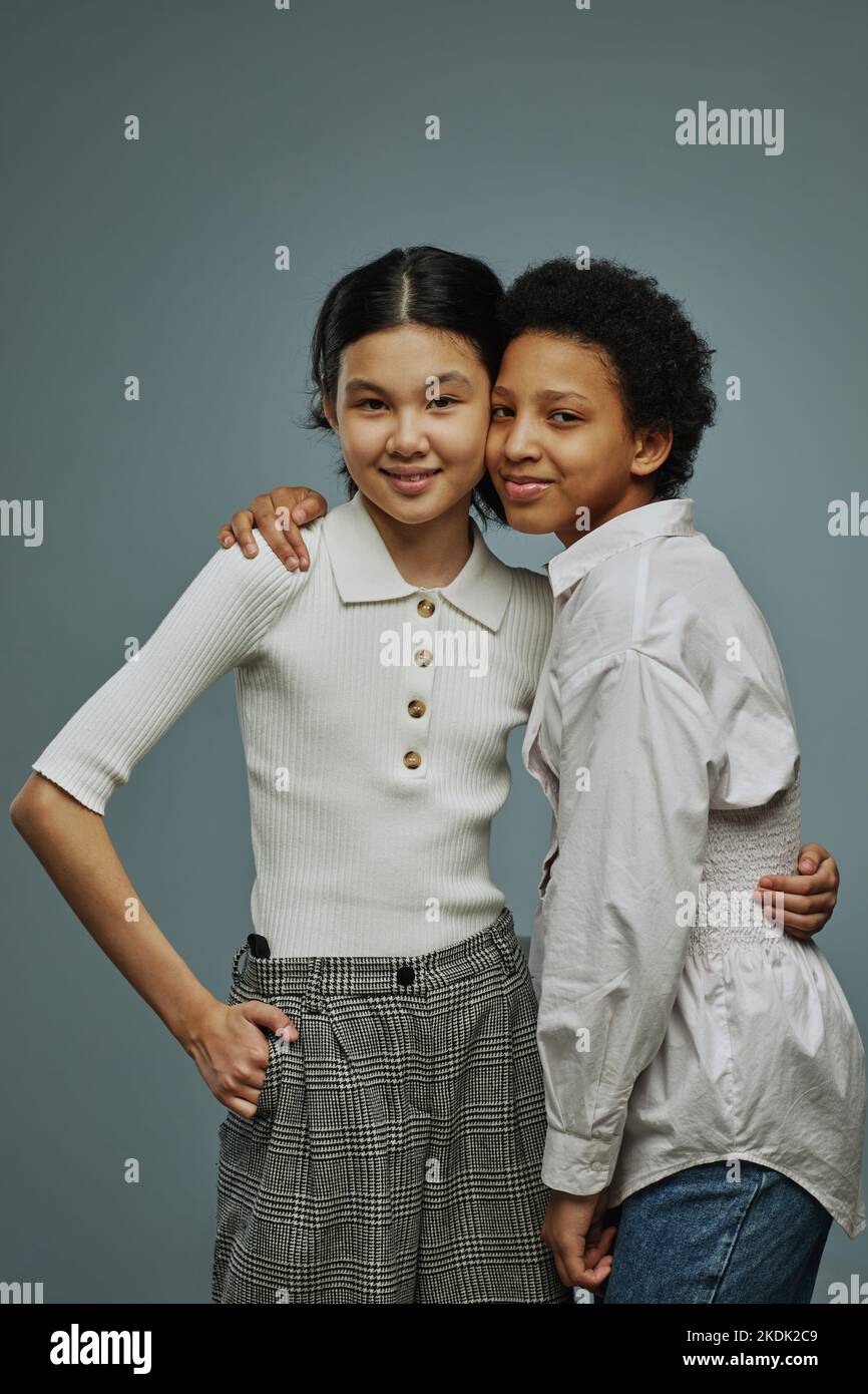 Two adorable intercultural schoolgirls in white casual tanktops embracing one another and looking at camera while posing over grey background Stock Photo