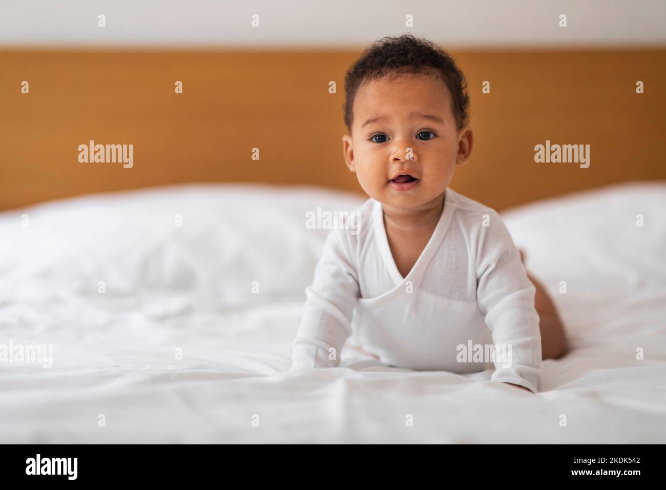 Cute Little African American Baby In White Bodysuit Lying On Bed Stock Photo