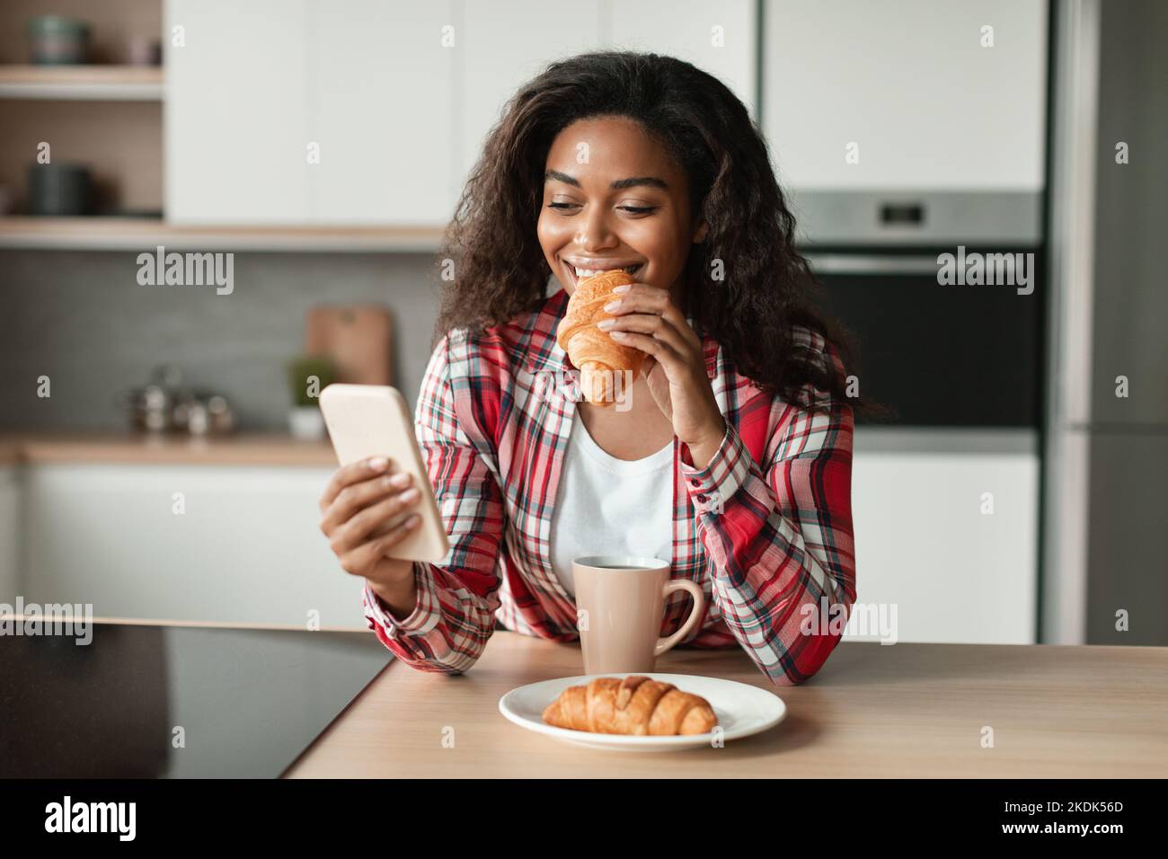 Glad black young woman in casual eat croissant, typing on phone, chatting, reading news and enjoy breakfast Stock Photo