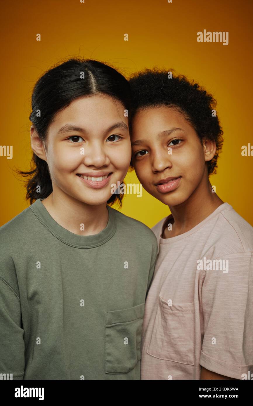Two cute friendly youthful girls of different ethnicities in t-shirts standing in front of camera while posing during photo session in studio Stock Photo
