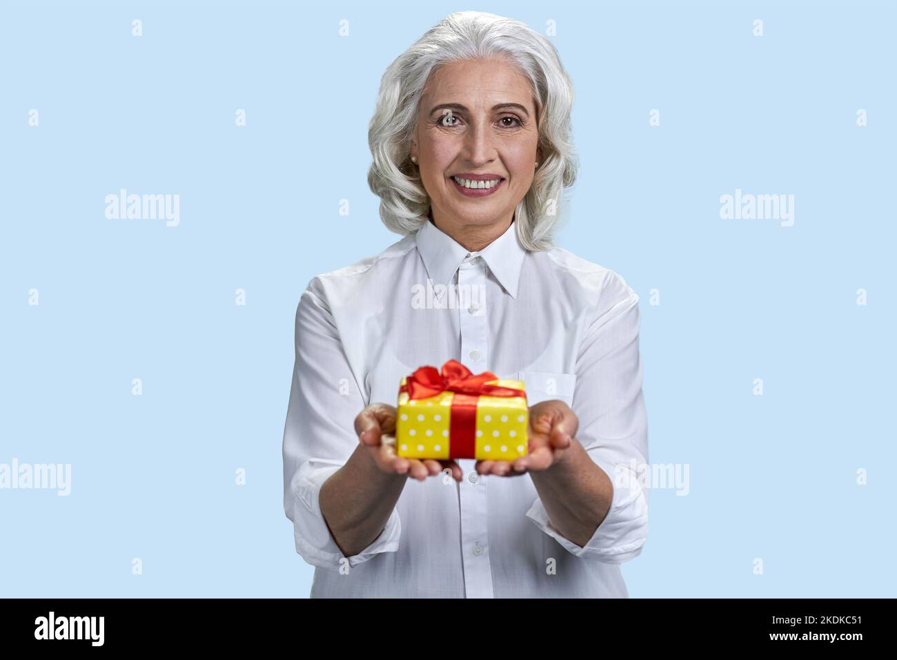 Portrait of happy gray-haired lady giving a yellow gift box. Happy old woman on pastel blue background. Stock Photo
