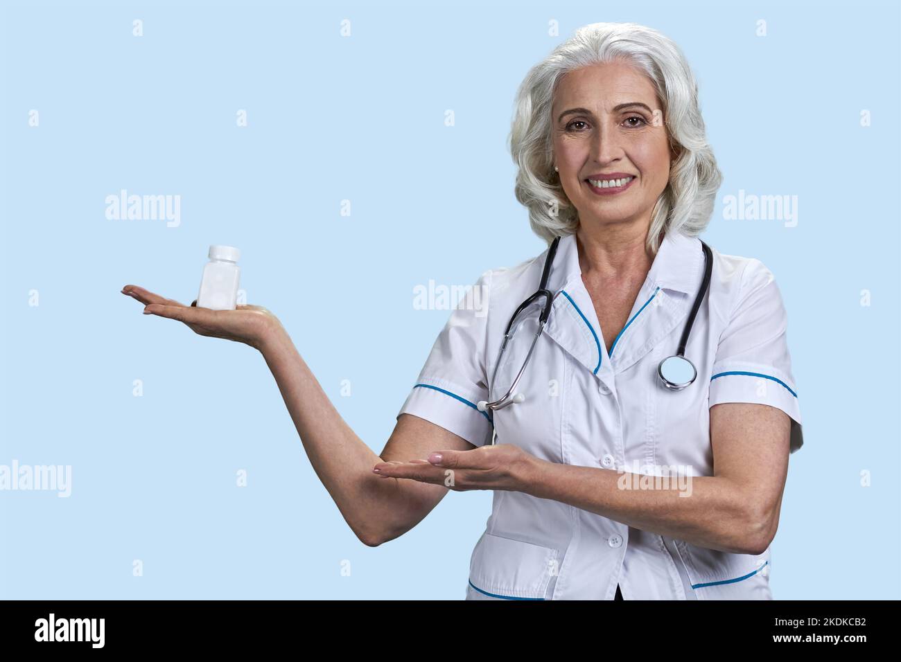 Portrait of senior aged woman advertising white medicine bottle. Gray-haired lady on blue background. Stock Photo