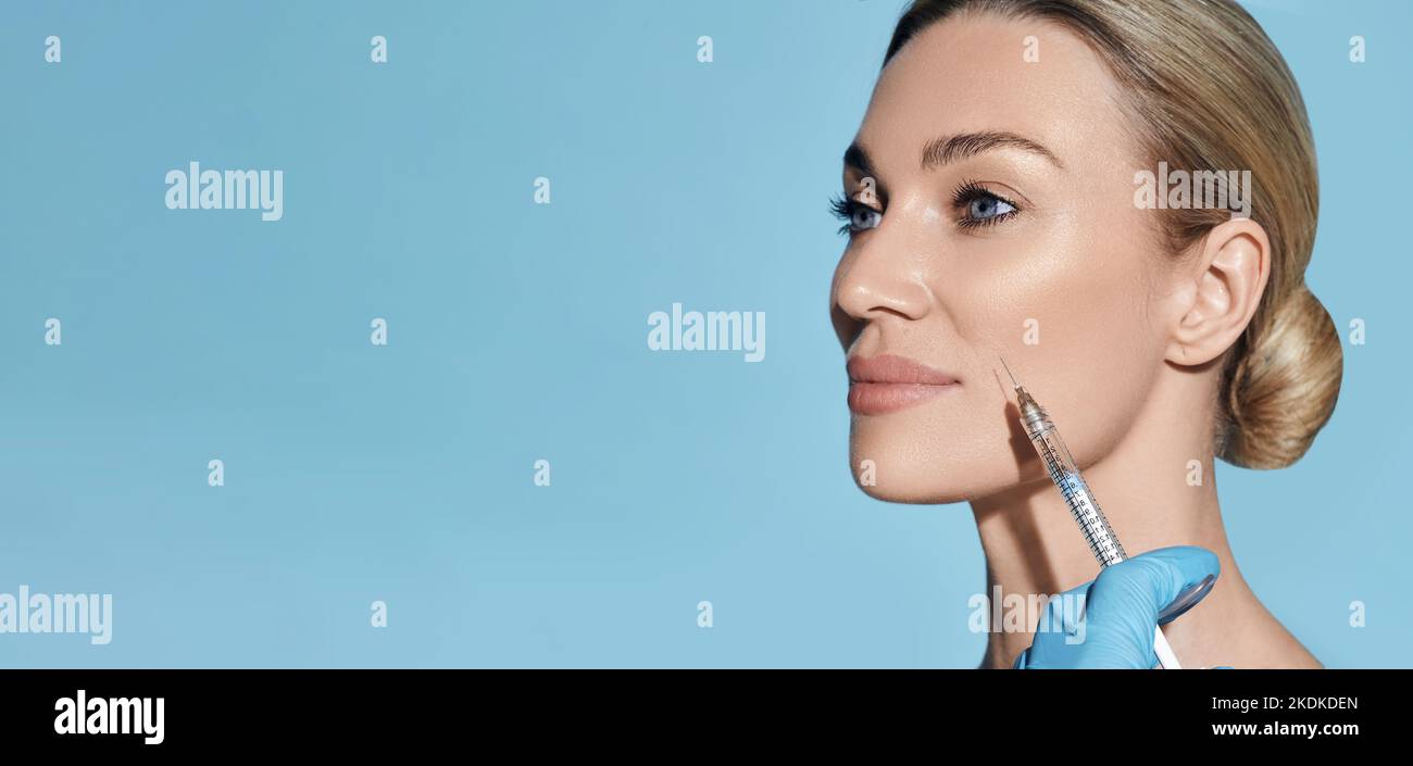 Beautician doing beauty injection into nasolabial folds on woman face with mimic wrinkles near mouth, on blue background. Botulinum toxin injections Stock Photo