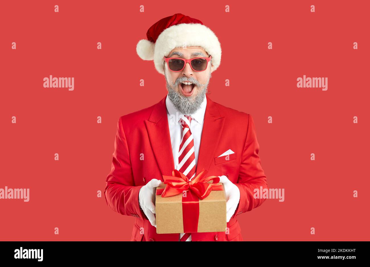 Old grey-haired happy showman in Santa style is giving a present, gift box on red background. Stock Photo