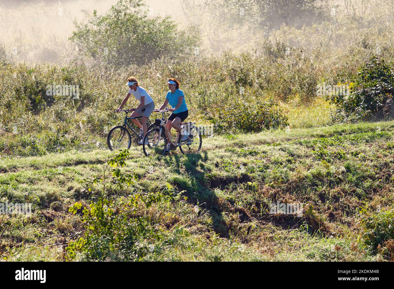 Two women riding bicycles along a mist-shrouded dike. Lower Mainland, B. C., Canada. Stock Photo