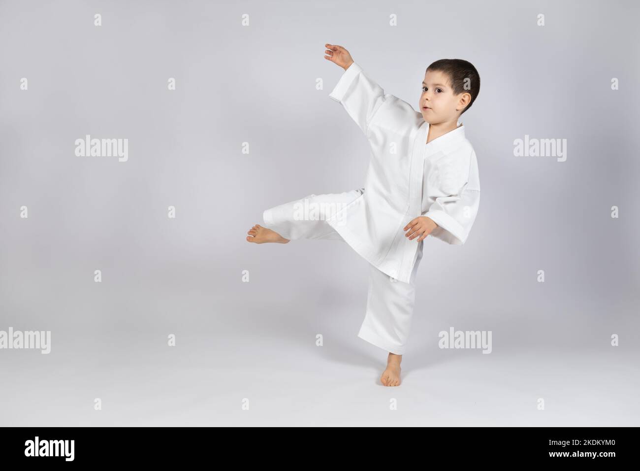 A little boy in a kimono practices karate on a white background, kicking forward. Stock Photo