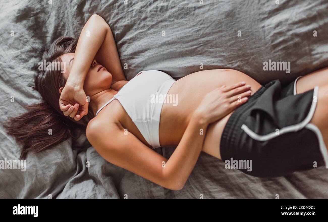 Tired pregnant woman sleeping on bed hiding face crying in depression or feeling morning sickness nausea. Could be used for Mental health, fatigue Stock Photo