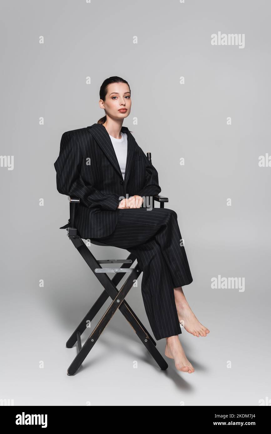 Full length of barefoot woman in striped suit sitting on folding chair on grey background Stock Photo
