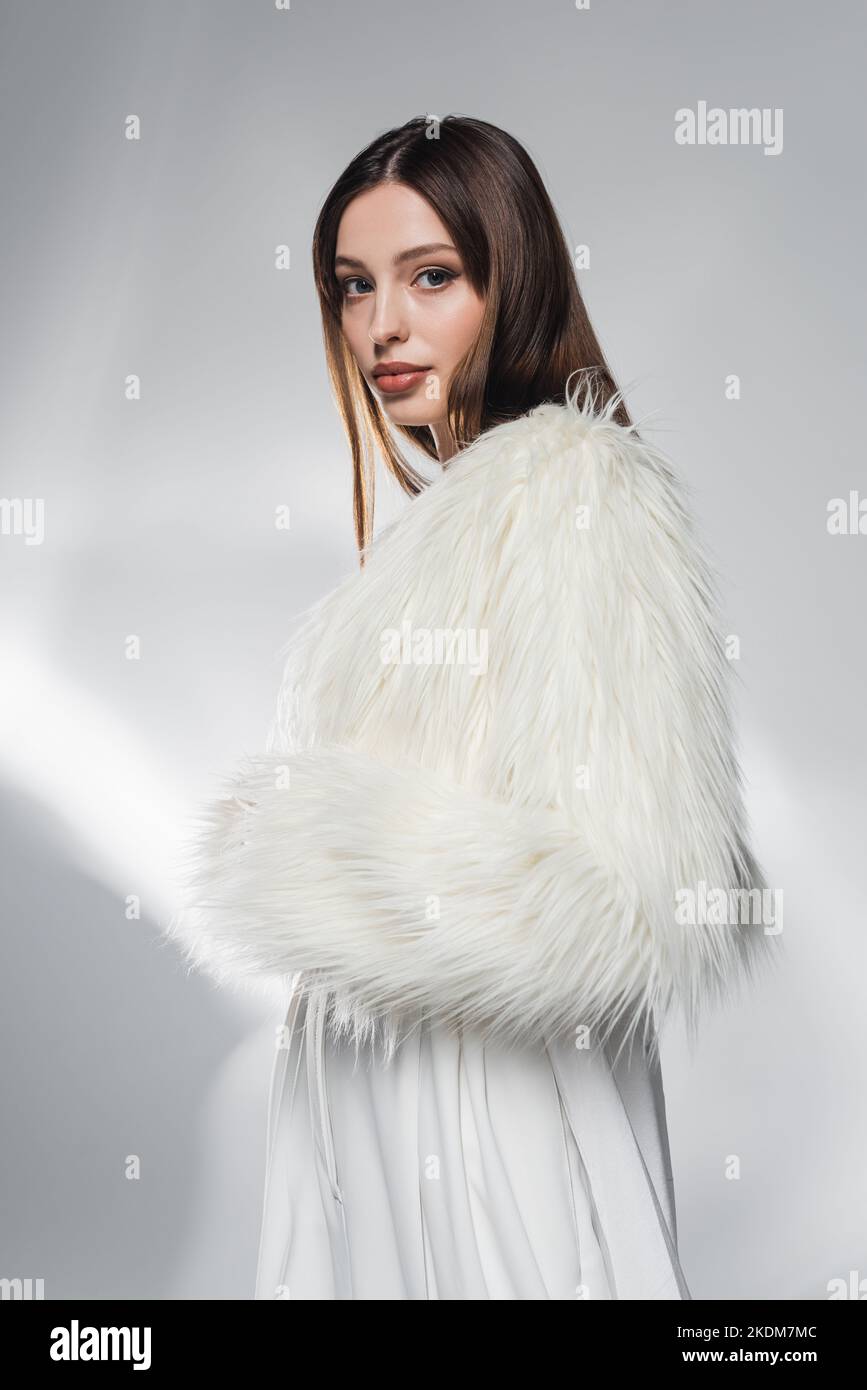 Stylish brunette woman in faux fur jacket looking at camera on abstract ...