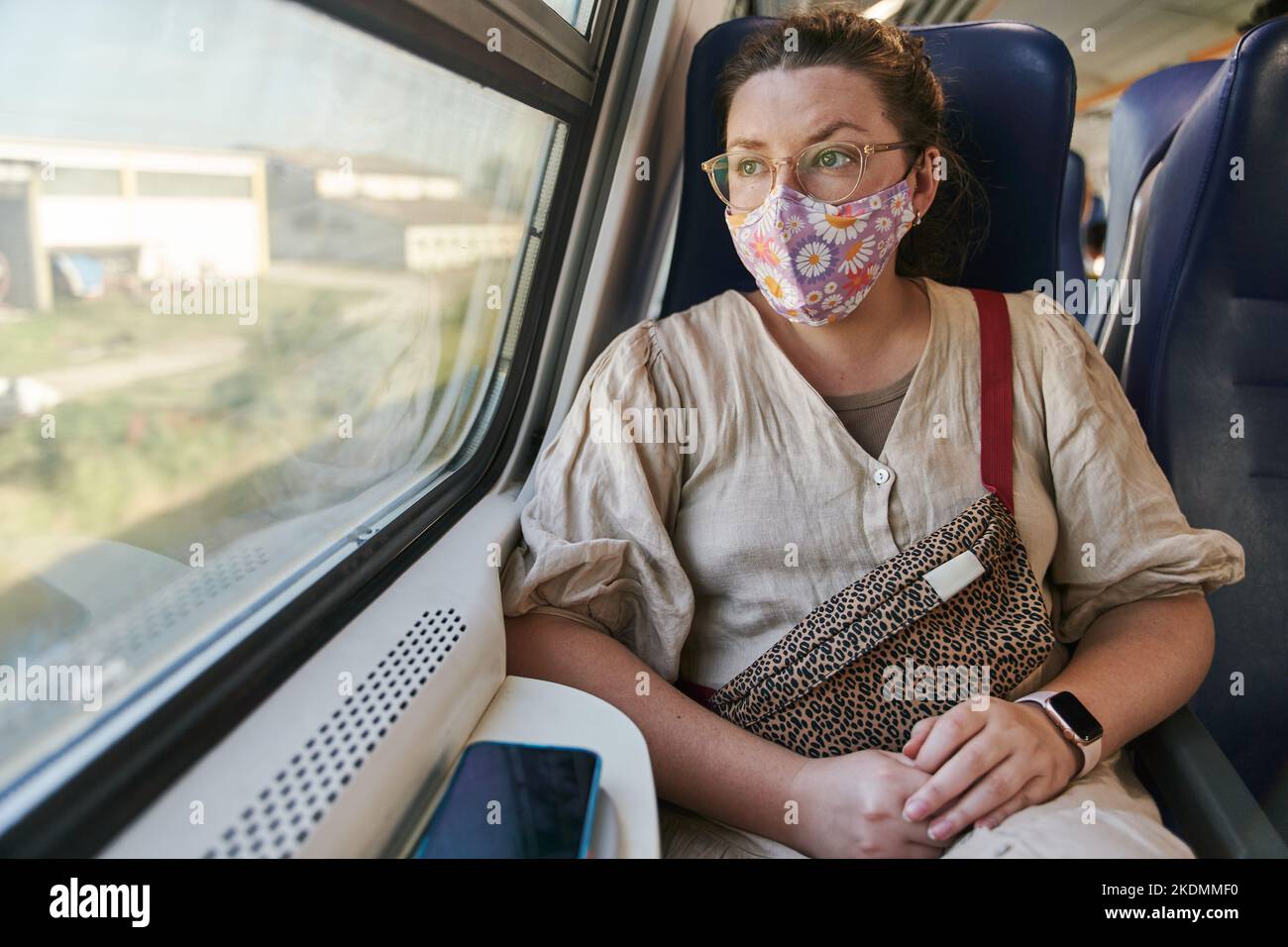 A girl in glasses and a medical mask riding a train and looking out the window Stock Photo