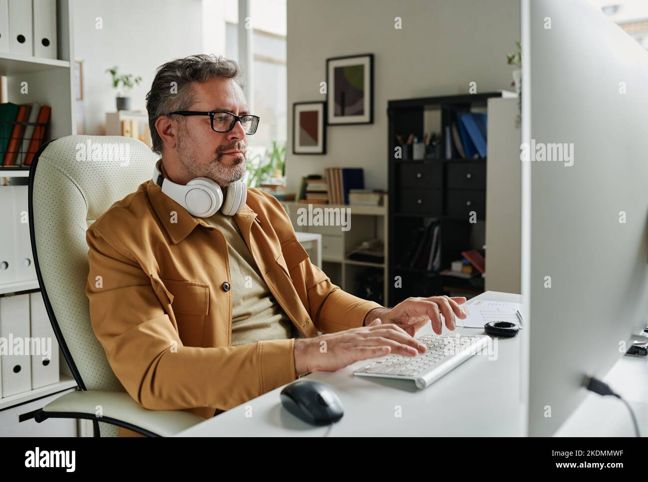 Serious grey haired programmer looking at computer screen while sitting by workplace in front of monitor and decoding data Stock Photo