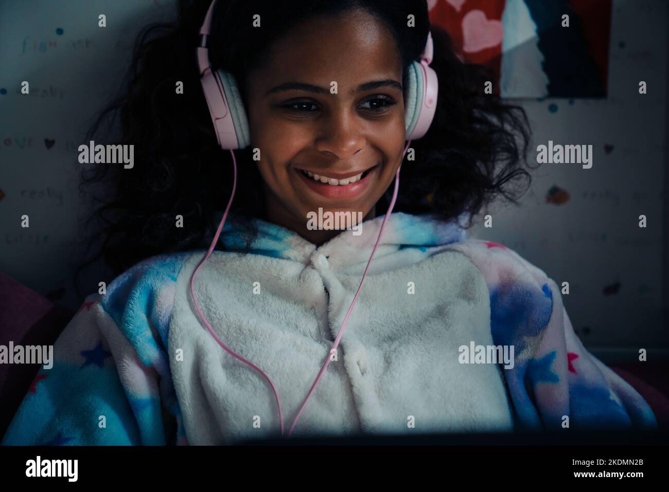 African happy smiling girl in pajamas and headphones looking on glowing screen at home Stock Photo