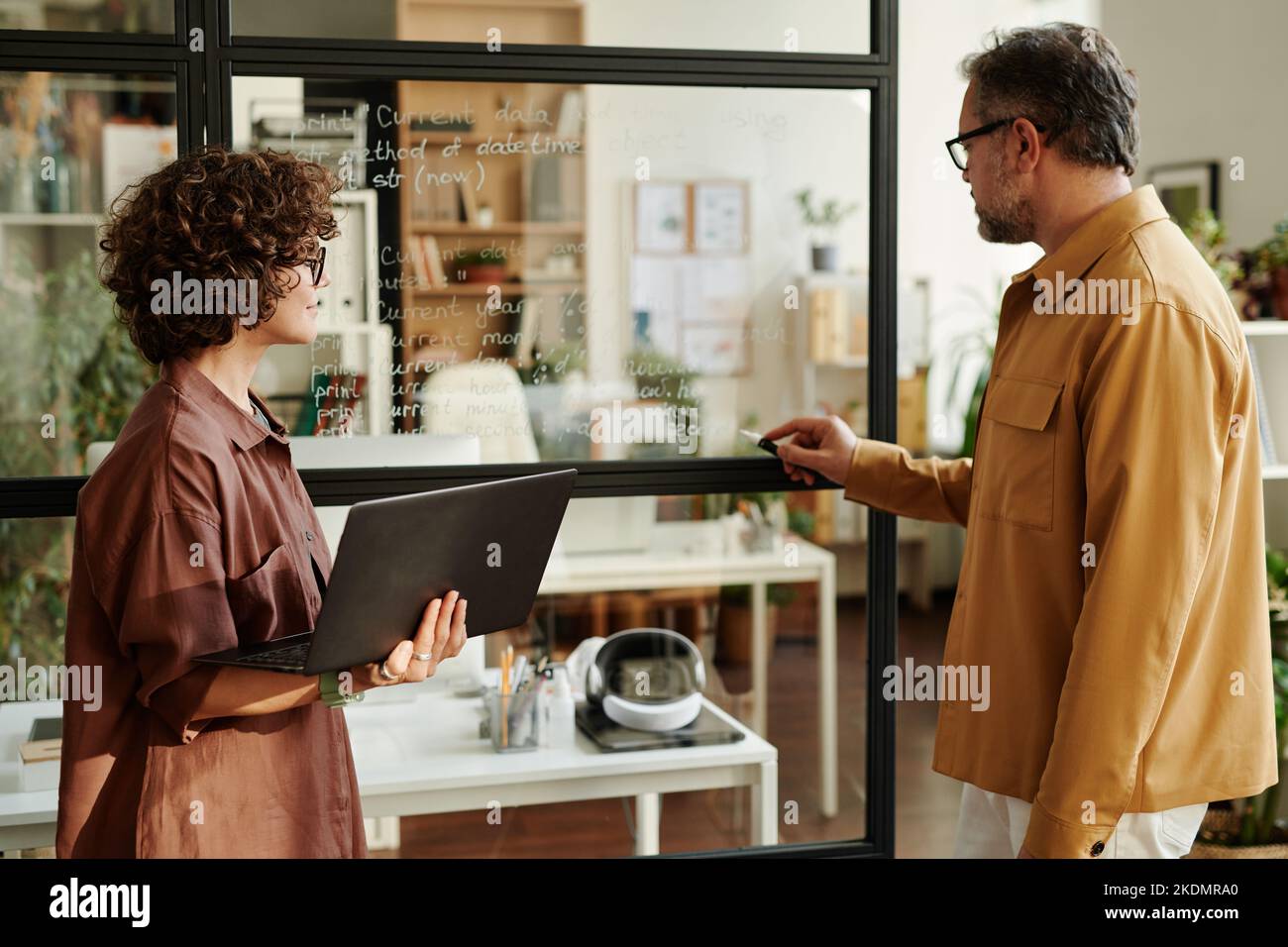 Confident mature man pointing at transparent board with notes while explaining them to young female colleague with laptop Stock Photo