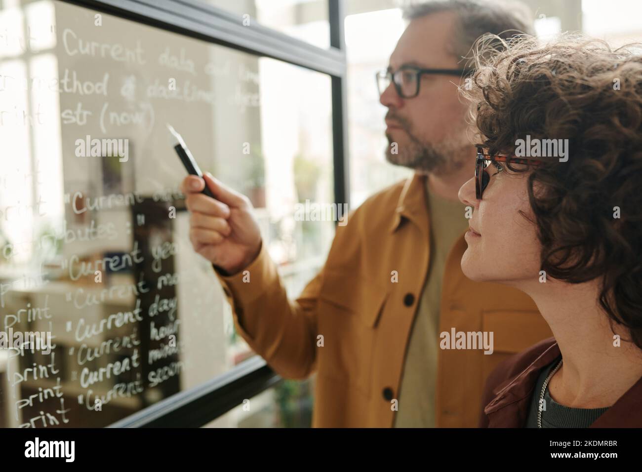 Selective focus on young businesswoman in eyeglasses listening to explanation of notes on transparent board written by mature colleague Stock Photo