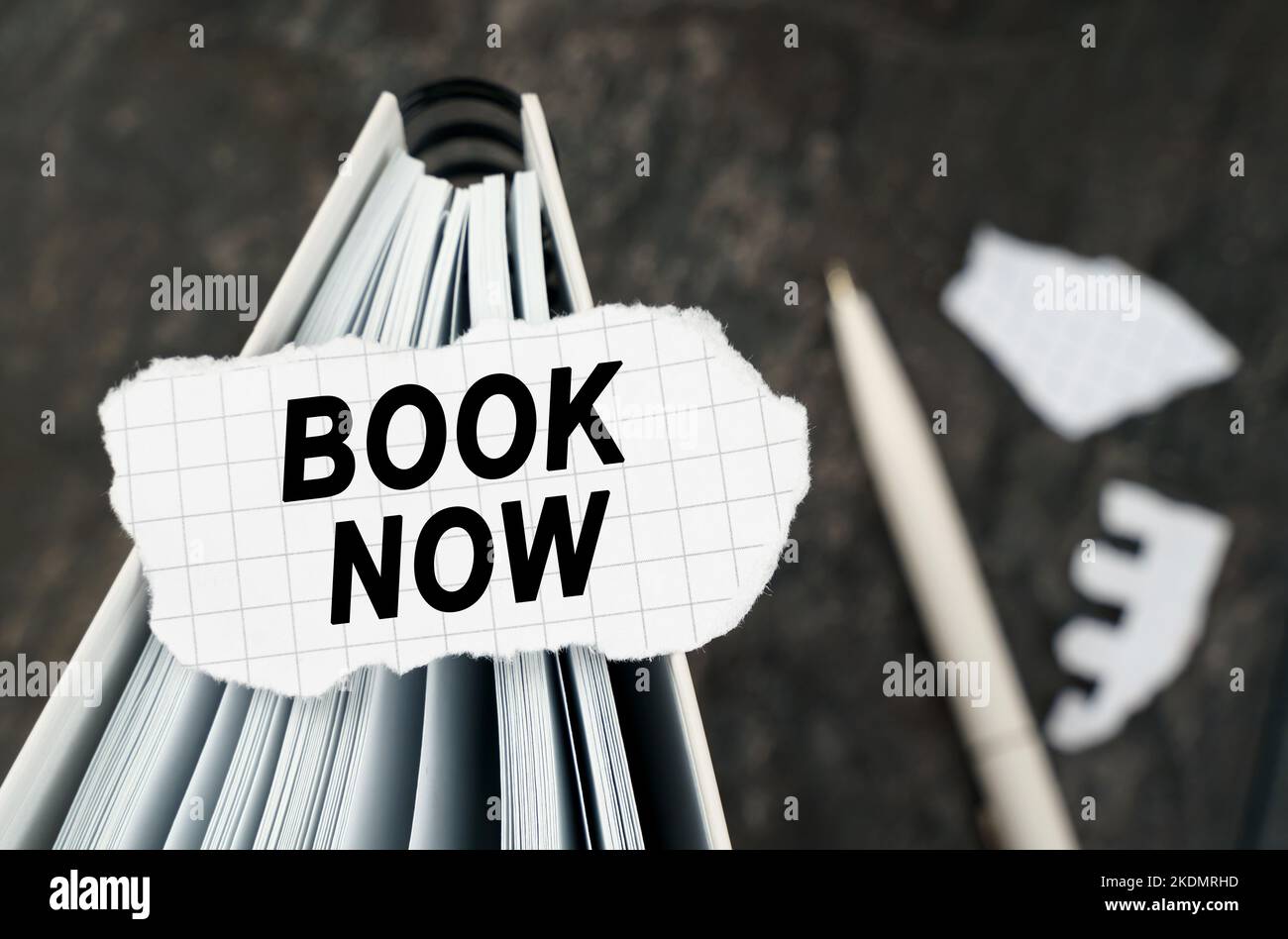 On the table is a notebook on which lies a piece of torn paper with the inscription - book now. The pen lies outside the sharpness zone. Stock Photo