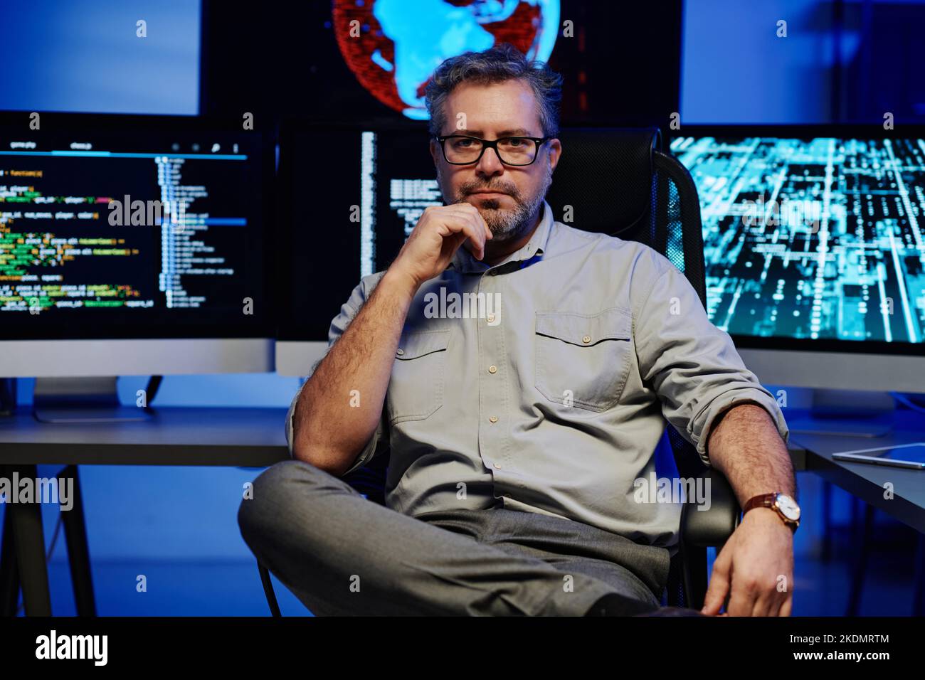 Serious mature male software developer in smart casualwear and eyeglasses sitting against computer monitors in server room Stock Photo