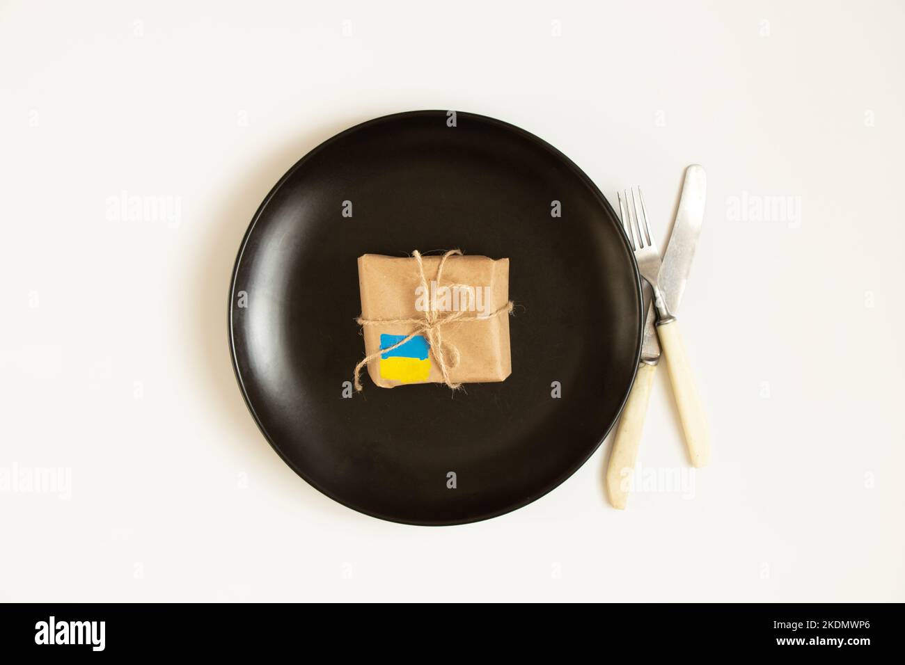 A box with a painted flag of Ukraine lies on a black plate and next to a knife with a fork on a white background, humanitarian aid for Ukraine, war Stock Photo