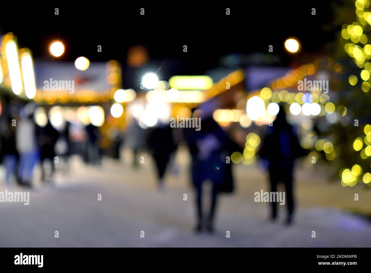 Blurred background. People walk in city square on winter night. Black silhouettes of people walking near houses decorated luminous illumination. White light bokeh blur spots from glowing house lights Stock Photo