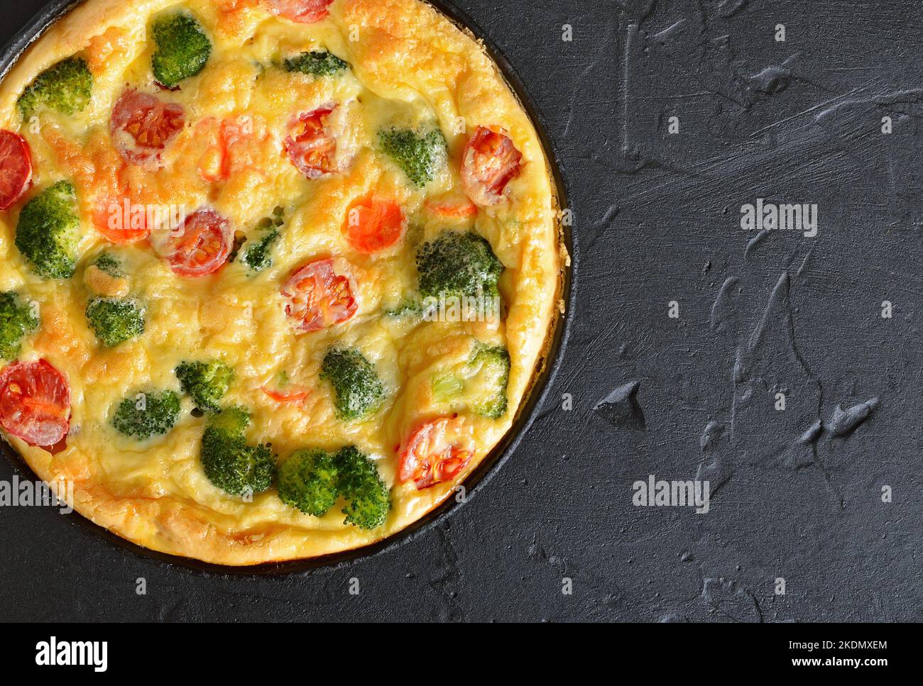 Frittata with broccoli and tomatoes in frying pan over black background. Top view, flat lay