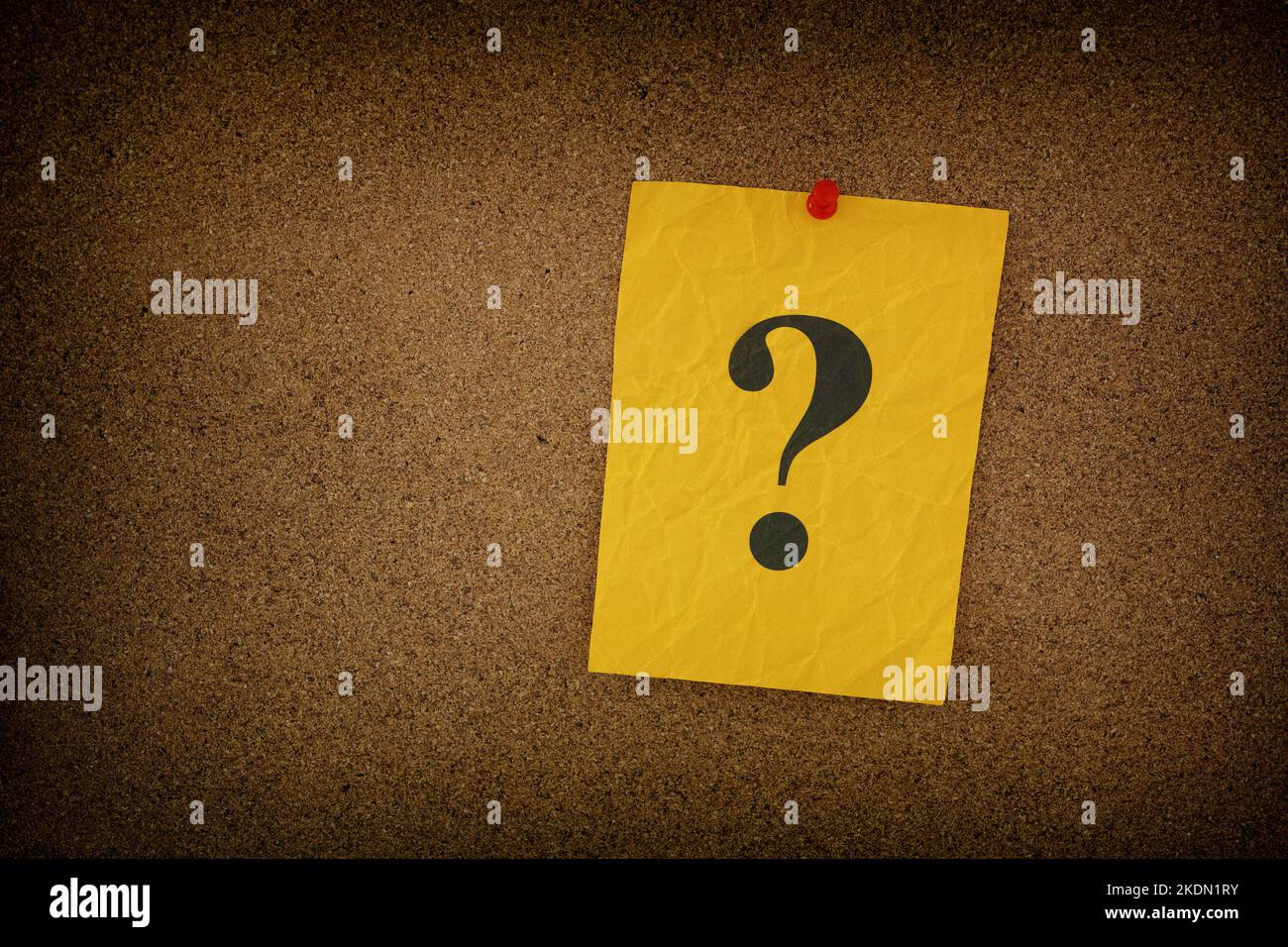 Yellow crumpled paper note with question marks on it pinned to a cork board. Closeup. Stock Photo