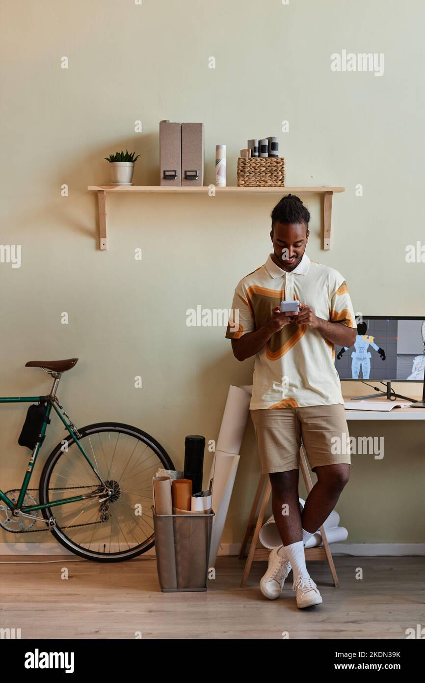 Vertical portrait of young black man as digital 3D designer using smartphone by workplace Stock Photo