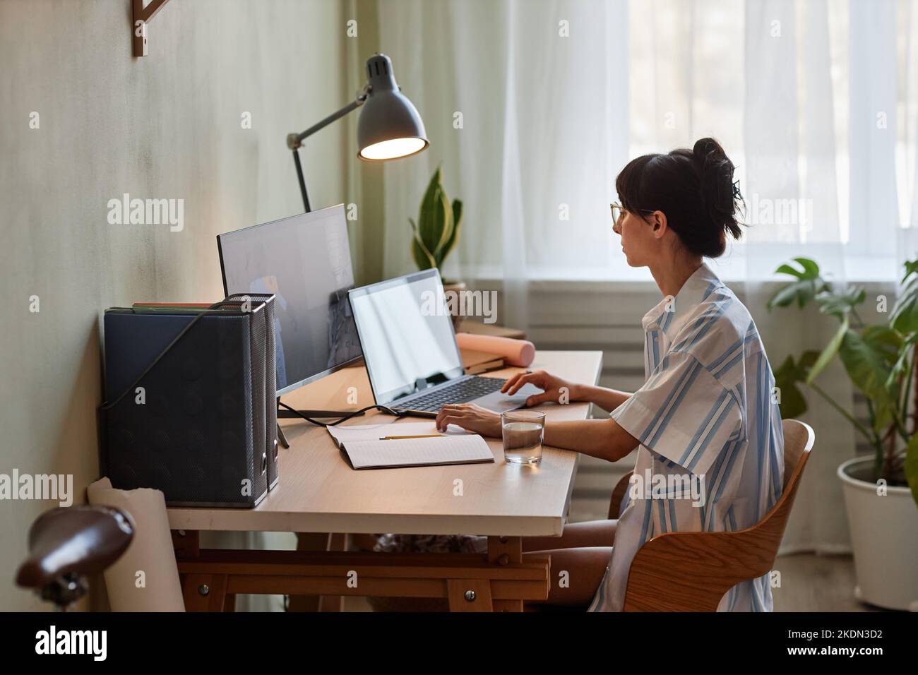 Side view portrait of young woman using laptop while working or studying at cozy home office workplace with dim lights Stock Photo