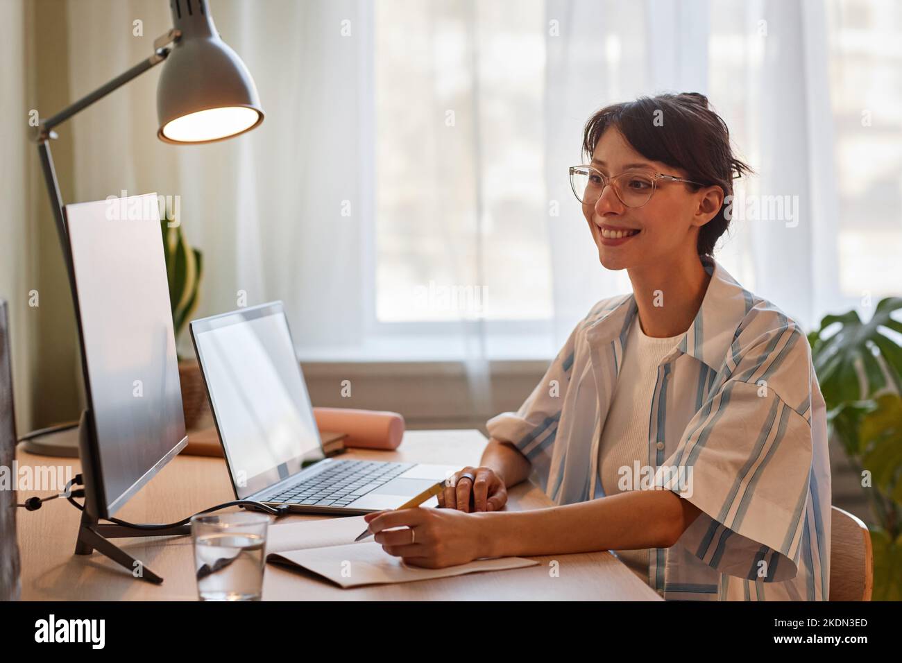 Portrait of smiling young woman enjoying online education class or work meeting at cozy home office workplace with dim lights Stock Photo
