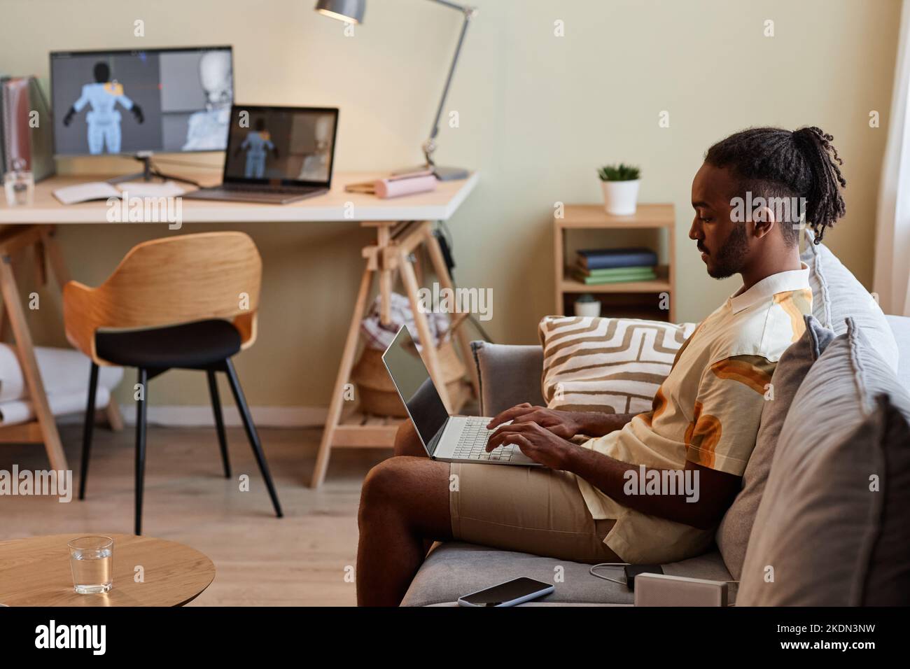 Side view portrait of creative black man using laptop while sitting on sofa in modern home interior, copy space Stock Photo