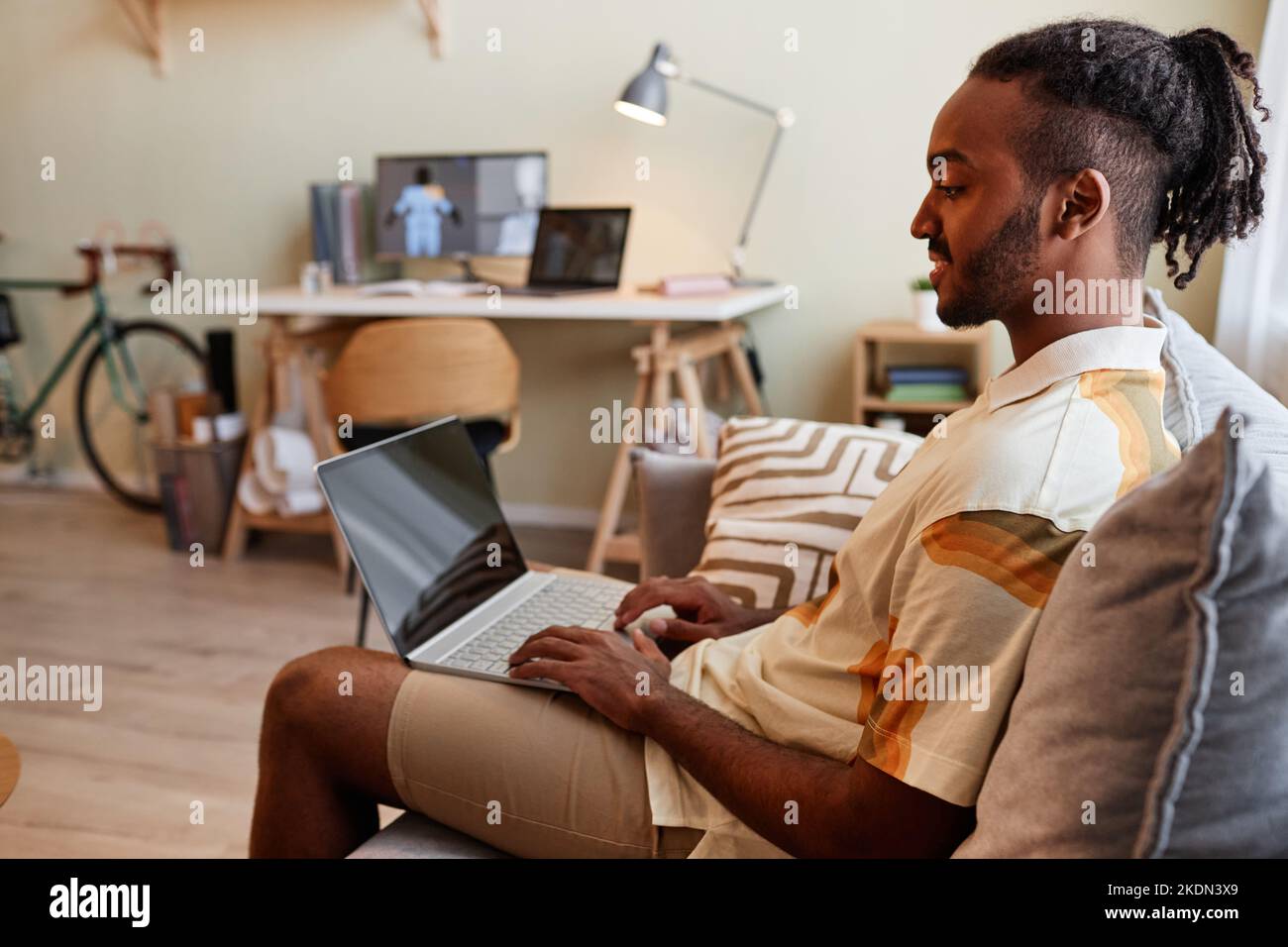 Side view portrait of creative black man using laptop while sitting on sofa and smiling at blank screen Stock Photo