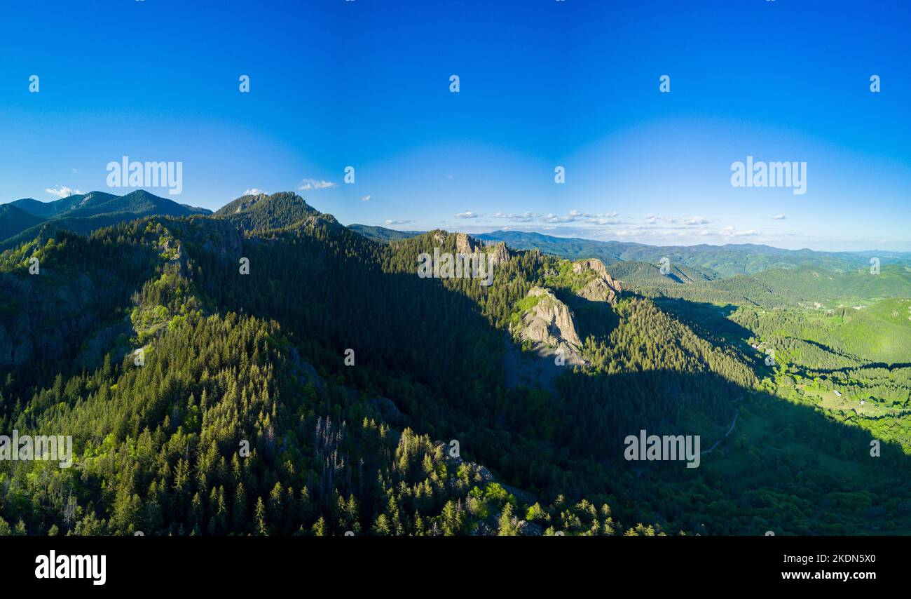 Rocky peak of high mountain with vegetation in valley of Rhodope mountains and forests against background of sky with large clouds. Panorama, top view Stock Photo