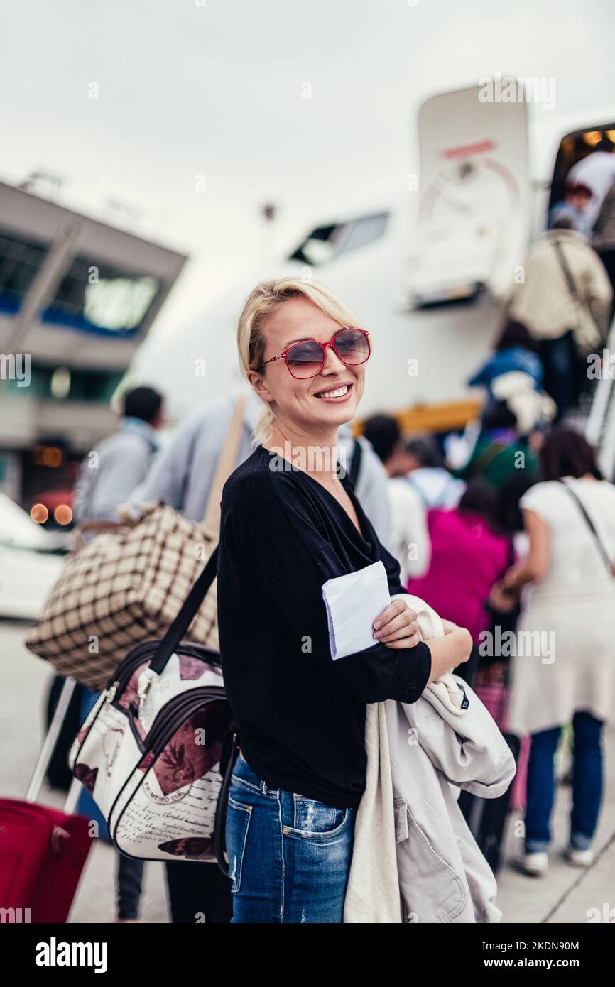 Cheerful woman holding carry on luggage queuing to board the commercial airplane. Stock Photo