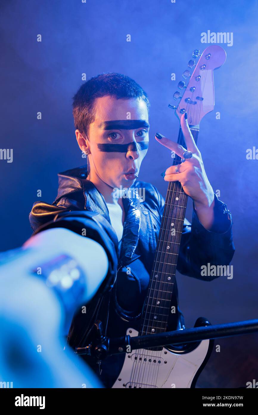 Female artist showing peace sign on camera, singing rock music and heavy metal alternative audio. Playing guitar and doing live performance in studio, doing victory symbol with instrument. Stock Photo