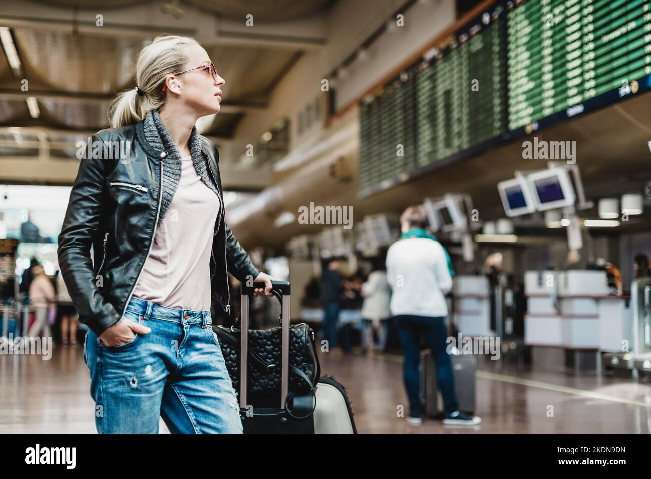 Casually dressed young stylish female traveller checking a departures board at the airport terminal hall in front of check in couters. Flight schedule display blured in the background. Focus on woman. Stock Photo