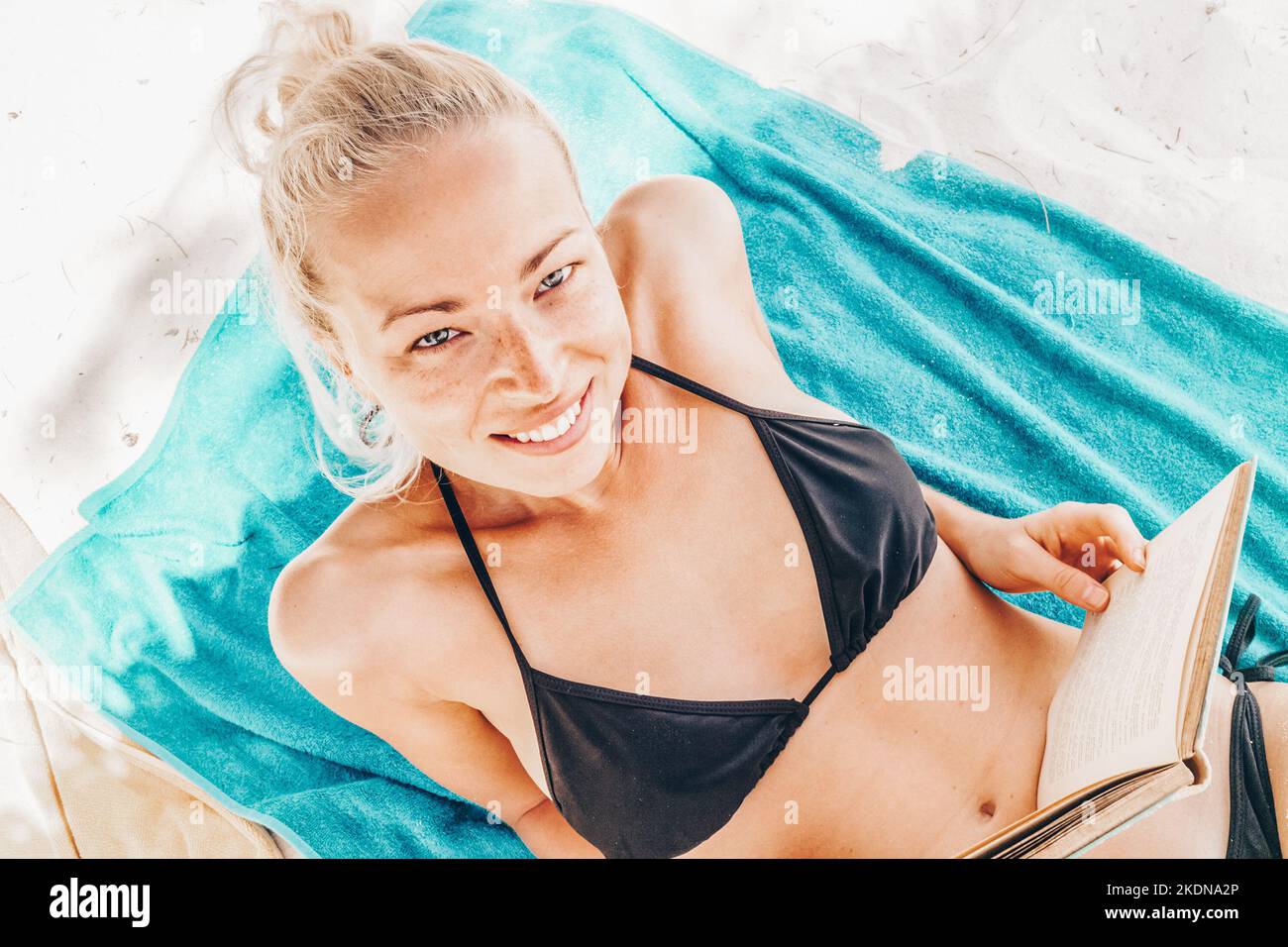 Young beautiful blonde caucasian woman in black bikini enjoying reading book on the beach while sunbathing. Stock Photo