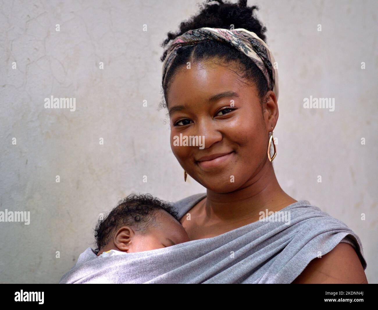 Relaxed smiling beautiful young black woman carries her sleeping newborn baby girl in an infant wrap sling and looks with confidence at viewer. Stock Photo