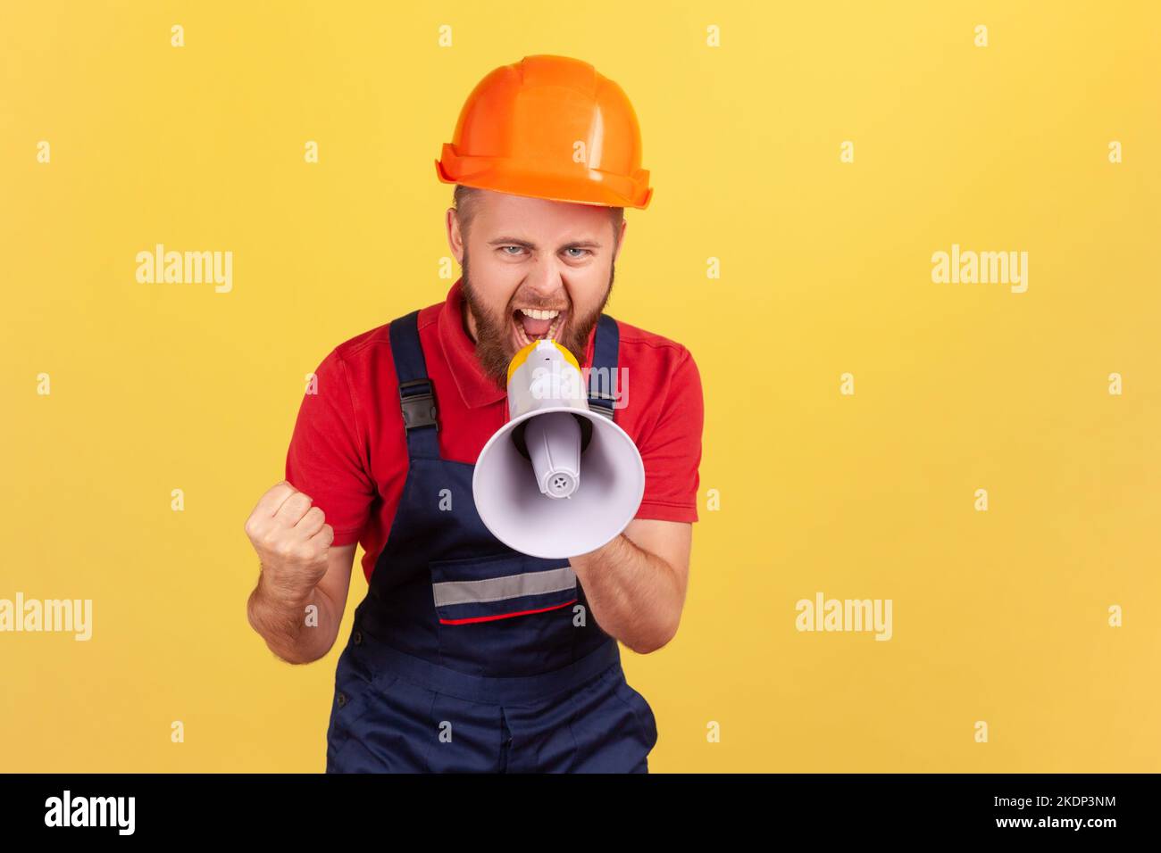 Portrait of angry aggressive bearded worker wearing protective helmet and blue overalls holding megaphone and screaming, protesting. Indoor studio shot isolated on yellow background. Stock Photo