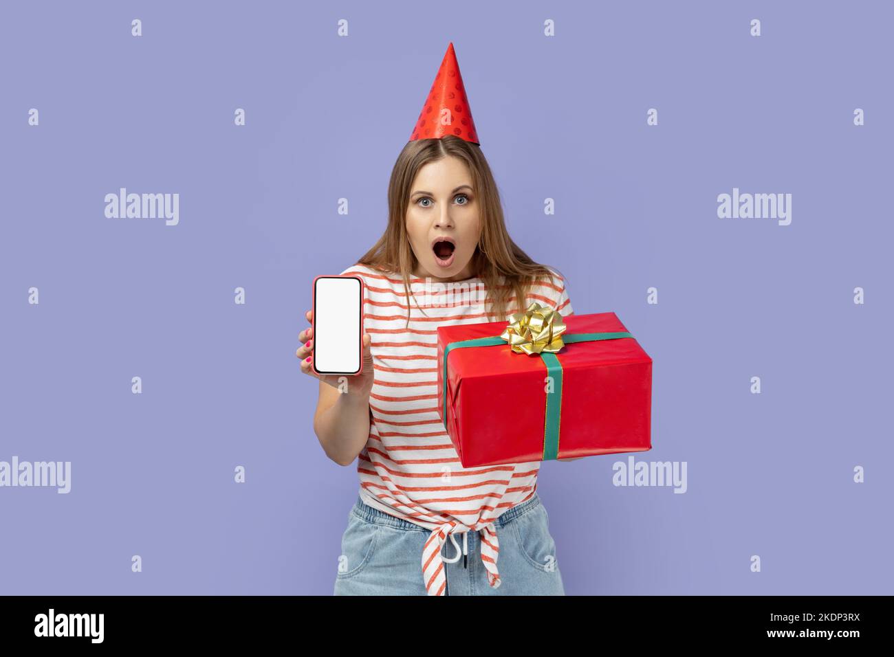 Portrait of shocked blond woman wearing striped T-shirt and party cone holding present box, holding smart phone with blank screen for promotional text. Indoor studio shot isolated on purple background Stock Photo
