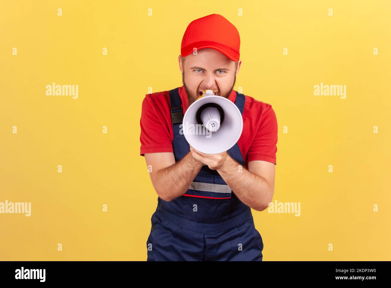 Portrait of angry worker man holding megaphone near mouth loudly speaking, screaming, making announcement, wearing blue uniform and red cap. Indoor studio shot isolated on yellow background. Stock Photo